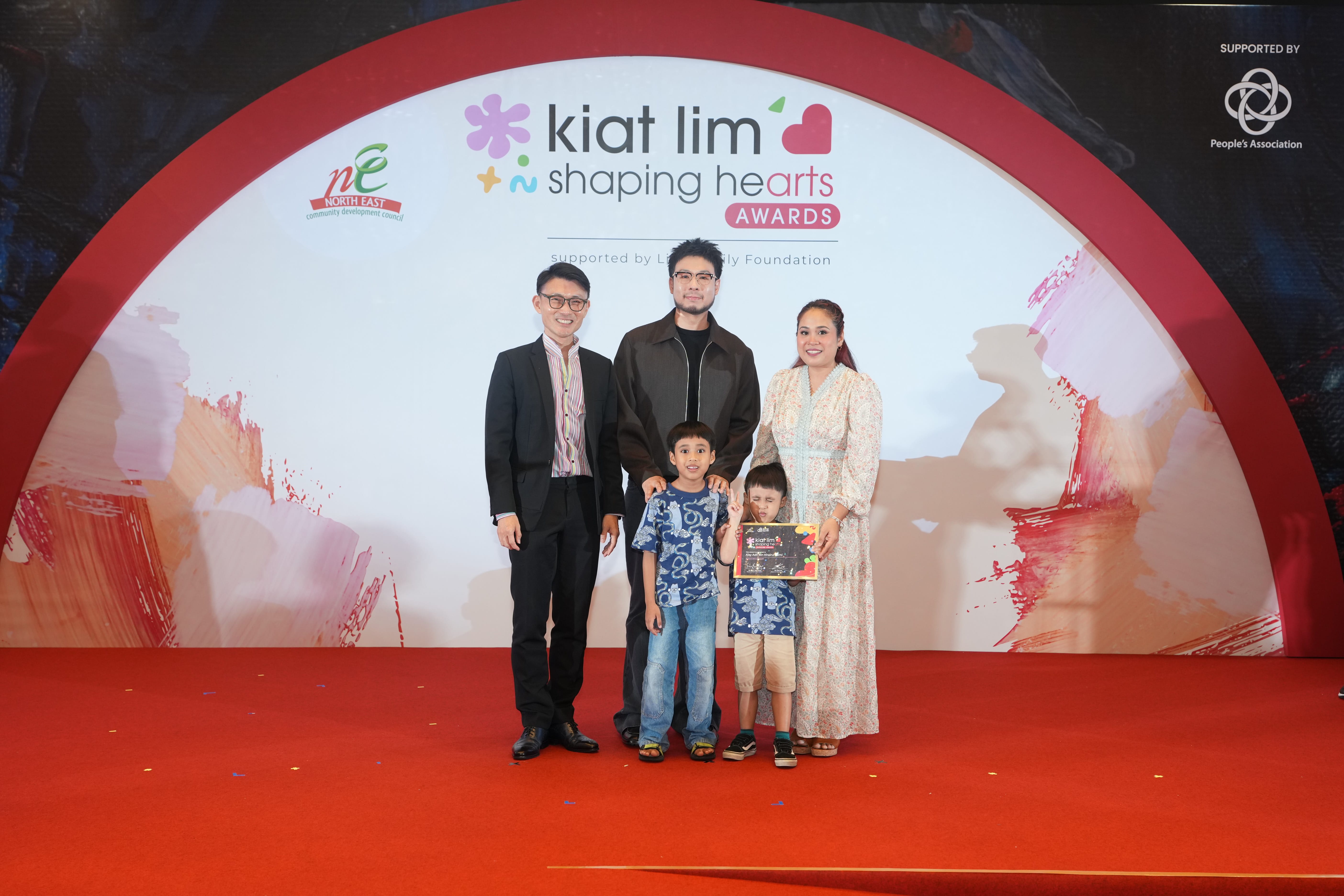Award recipient standing on stage with the presenter, holding their certificate and posing for a group photo during the award ceremony, with a backdrop reading ‘Kiat Lim-Shaping Hearts Awards'.